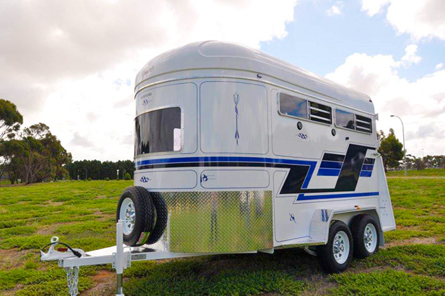 White travel trailer with blue and black graphics on a grassy field; tandem axle and front spare tire with diamond-plate panel.
