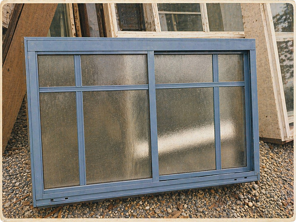 Blue-framed double-window pane leaning on a gravel surface with other window frames in the background.